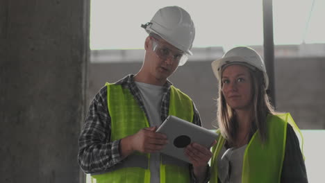Zwei-Ingenieure,-Ein-Mann-Und-Eine-Frau-In-Weißen-Helmen-Mit-Einem-Tablet-Computer-Auf-Der-Baustelle-Verurteilen-Den-Bauplan-Des-Gebäudes